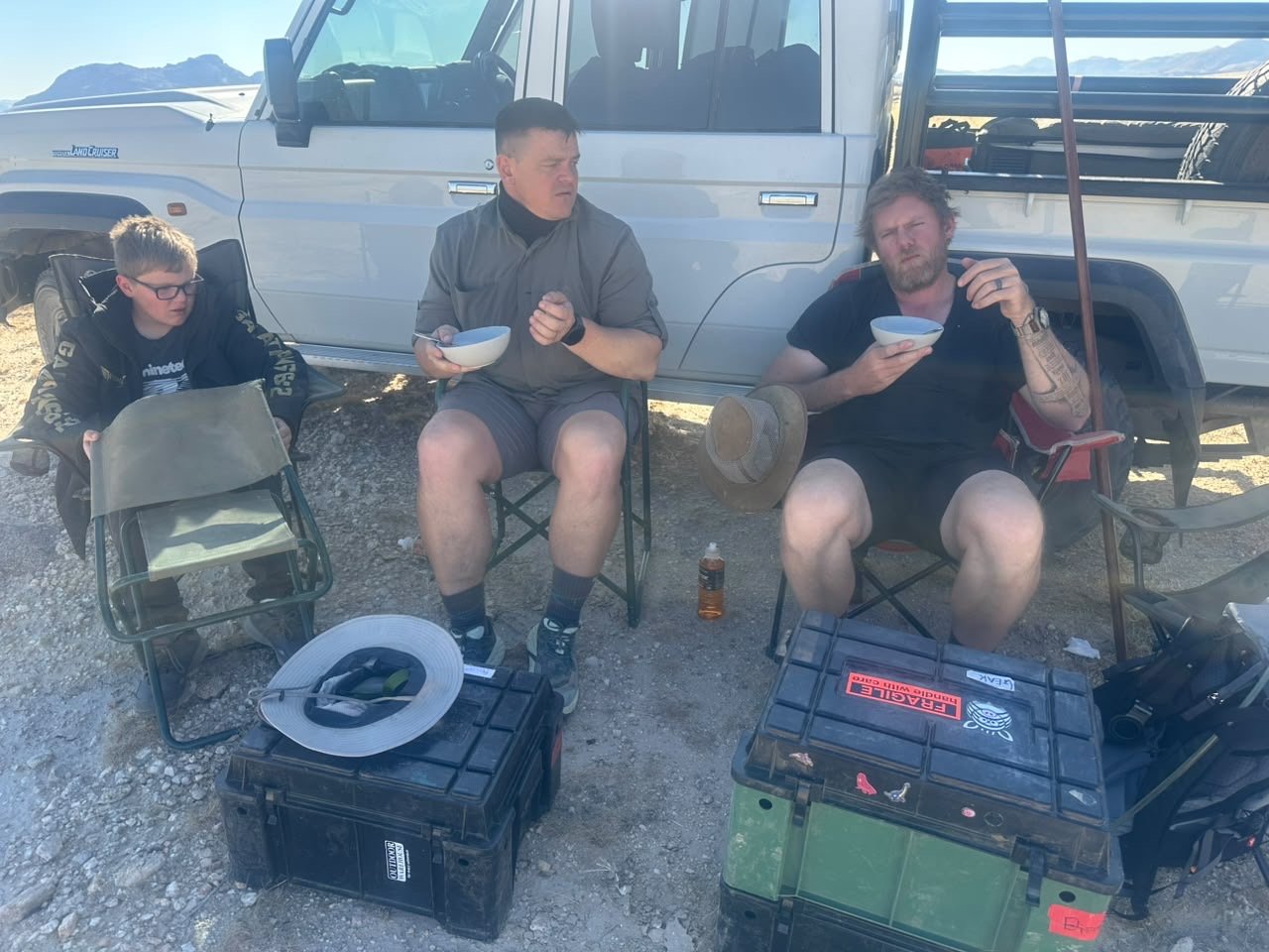 Companions taking a meal break beside a vehicle on an outdoor journey